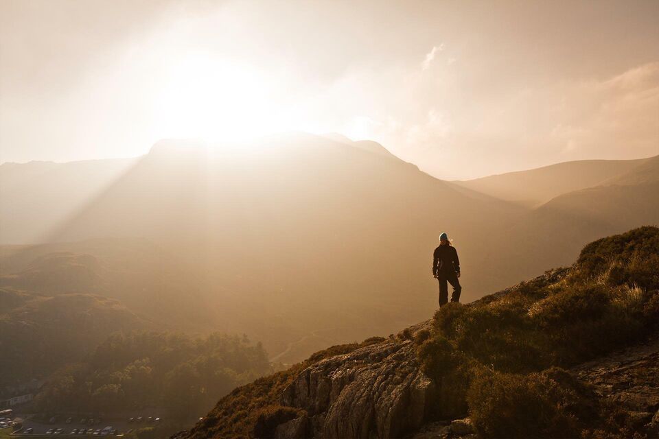 A mountainous landscape vista with the setting sun silhouetting a lone hiker on a ridge.